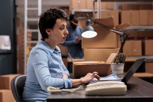 Woman manager looking at cargo stock on laptop computer while checking trendy blouse status before start preparing customers packages. Employee working at warehouse delivery department