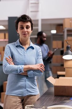 Woman supervisor standing in warehouse with arm crossed after finishing packing clients orders preparing packages for delivery. Diverse team working in delivery department in storehouse