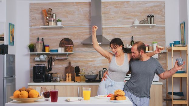 Carefree happy and joyful couple dancing and singing in kitchen in synny morning. Cheerful husband and wife laughing, singing, dancing listening musing, living happy and worry free. Positive people