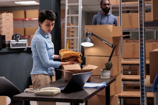 Employee putting trendy clothes in cardboard boxes preparing clients orders for delivery, checking shipping detalies on laptop computer in storehouse, Worker analyzing transportation logistics