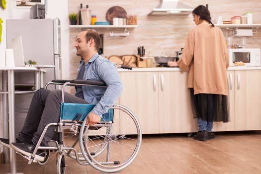 Disabled guy in wheelchair on video call in kitchen while wife cooking meal. Disabled paralyzed handicapped man with walking disability integrating after an accident.