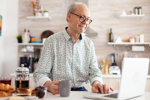 Mature man typing on laptop in kitchen during breakfast and driking coffee. Elderly retired person working from home, telecommuting using remote internet job online communication on modern