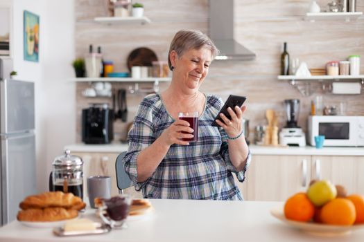 Senior woman using mobile gadget in the kitchen drinking aromatic tea in the morning during breakfast. Authentic elderly person searching on modern smartphone internet technology, senior leisure time