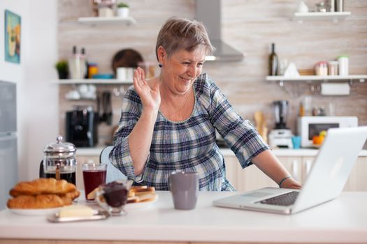 Retired woman waving during online meeting with family on video call using laptop in kitchen having breakfast. Elderly person using internet online chat tech, pc webcam for virtual conference call