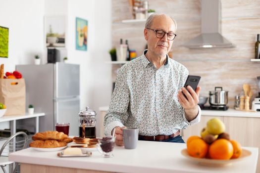 Happy old man surfing on social media using smartphone during breakfast sitting in kitchen smiling. Authentic portrait of retired senior enjoying modern internet online technology, searching, browsing