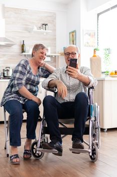 Smiling senior woman and her disabled husband in wheelchair using smartphone in kitchen, surfing on social media in the morning. paralyzedhandicapped old man using modern communication techonolgy.