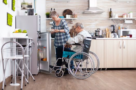 Senior woman preparing breakfast for handicapped husband taking eggs carton from refrigerator , living with man with walking disabilities. Disabled senior male in wheelchairhelping his wife in kitchen.