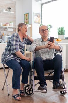 Senior woman and disabled husband in wheelchair surfing on internet using smartphone in kitchen. paralyzedhandicapped old elderly man using modern communication techonolgy.