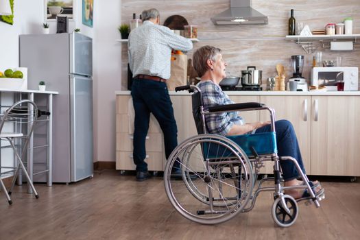 Paralysed senior woman in wheelchair feeling unhappy sitting in the middle of kitchen while husband unpacking grocery bag. Old handicapped lady after injury and rehab, depressed invalid full of sorrow