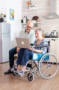 Handicapped senior woman in wheelchair and her husband searching on laptop, surfing on social media sitting in kitchen in the morning. paralyzeddisabled old person having a online conference.