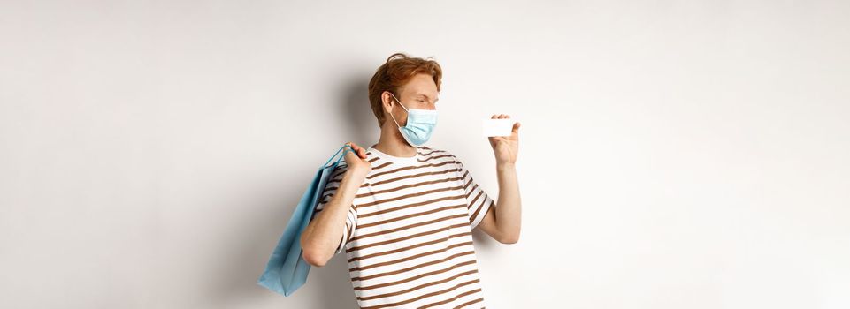 Concept of covid-19 and shopping. Happy young shopper in face mask holding paper bag and showing plastic credit card, buying with discounts, white background.