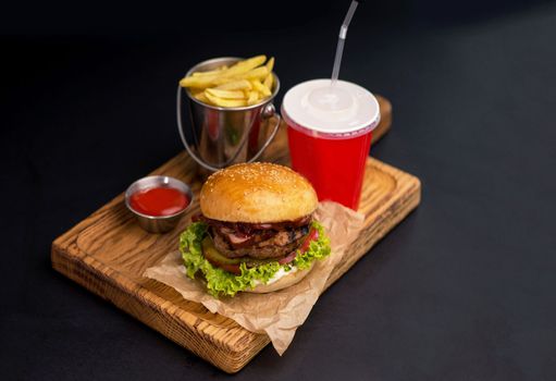 Classic burgers with becon, meat, cheese, onion, tomato and cheese and integral bun. On textured black board. Close up. Fried potatoes and glass of cola.