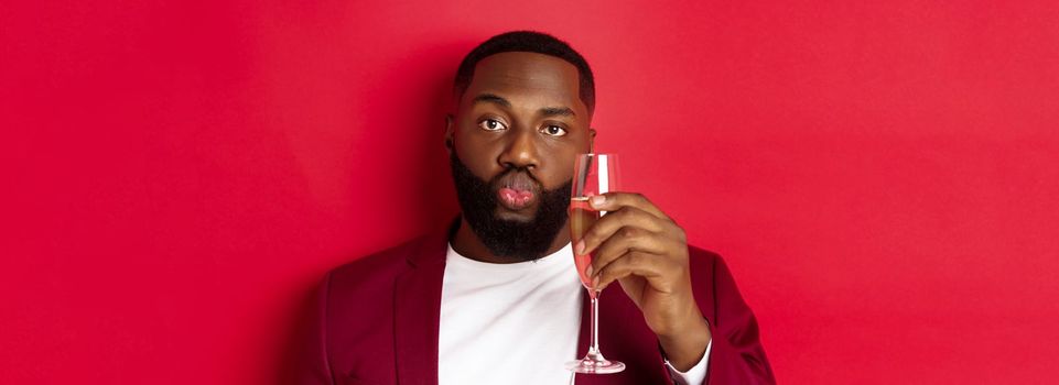 Close-up of funny Black man tasting champagne from glass, looking silly and pucker lips, celebrating new year, standing against red background.