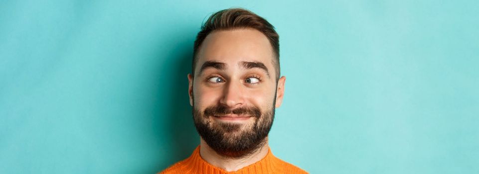 Headshot of funny caucasian man making faces, squinting and smiling silly, standing over light blue background.