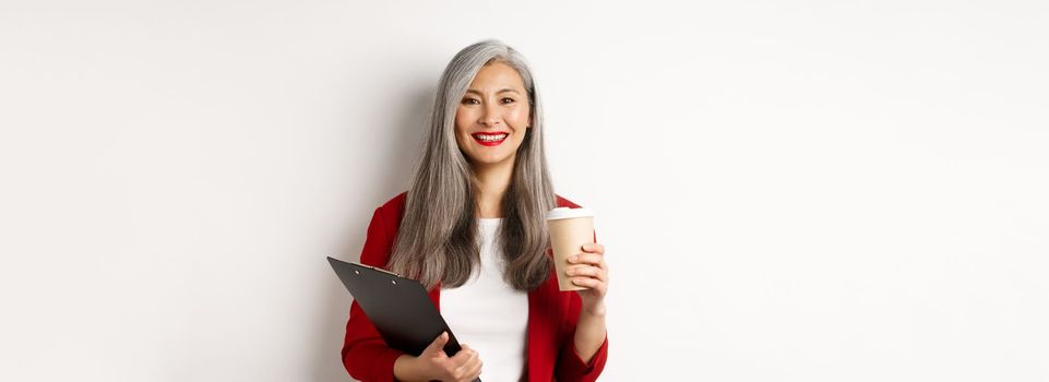 Successful asian businesswoman drinking coffee and smiling, standing with clipboard, wearing red blazer, white background.