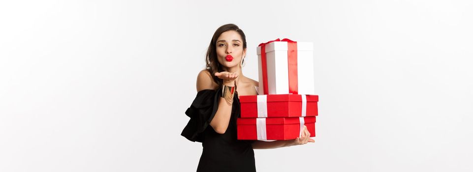 Celebration and christmas holidays concept. Pretty woman in elegant black dress holding presents, sending air kiss at camera, standing over white background.