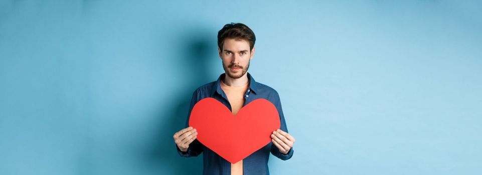 Young man looking for soulmate on valentines day, holding big red heart and looking at camera, standing over blue background.