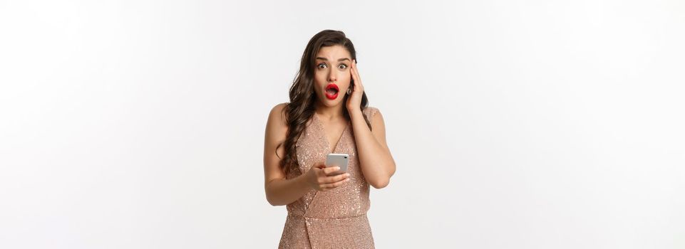 Holidays, online shopping concept. Surprised woman using mobile phone and looking amazed, wearing party dress, standing over white background.