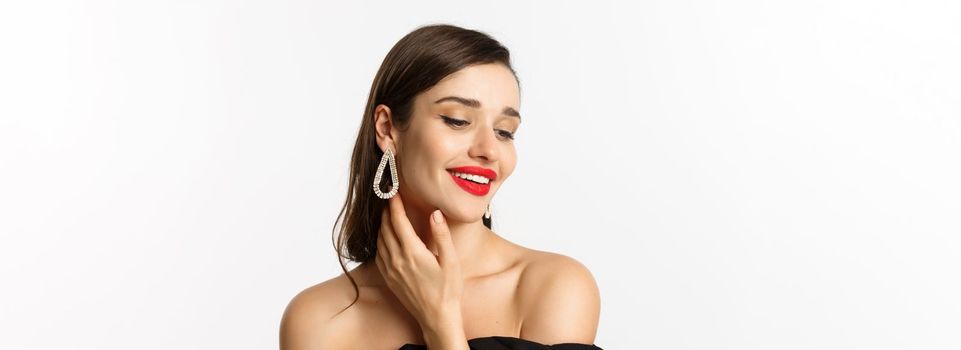 Fashion and beauty concept. Close-up of tender woman in black dress and earrings, gently touching face and smiling, looking down coquettish, standing over white background.