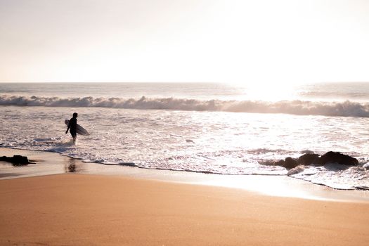 silhouette of an unrecognizable surfer walking along the shore of the beach with his board under his arm at sunset, concept of leisure and relax, copy space for text