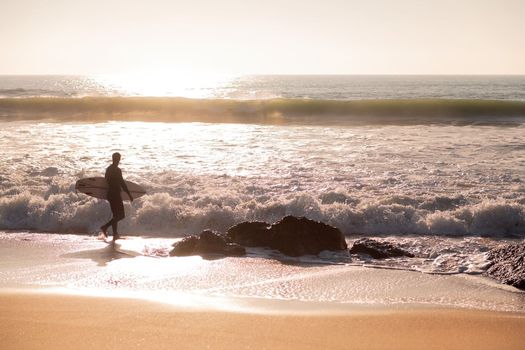 silhouette of a surfer walking along the shore of the beach with his board under his arm at sunset, concept of leisure and relax, copy space for text