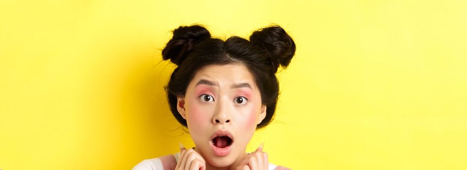 Head shot of shocked asian girl with hairbuns and glamour makeup, open mouth and looking startled at camera, standing against yellow background.