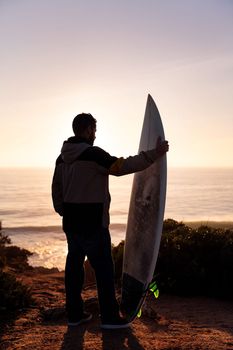 vertical photo of the silhouette of a young man in jacket looking at the sea at sunset next to his surfboard, leisure and hobbies concept, copy space for text