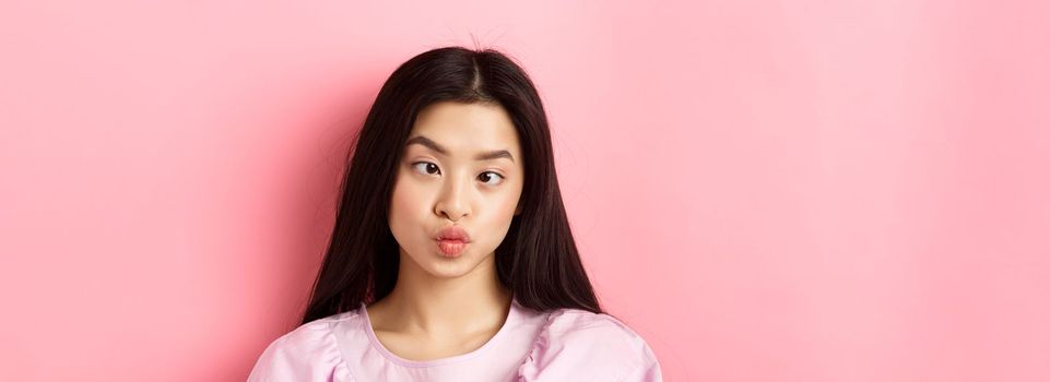 Close-up portrait of funny asian woman squinting eyes and making silly faces, standing against pink background.