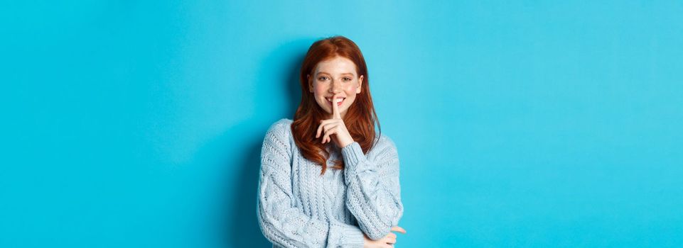 Pretty redhead teenager hushing and smiling, telling a secret, standing in sweater against blue background.