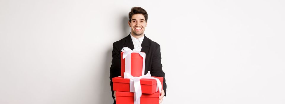 Portrait of handsome bearded man in trendy suit, holding gifts for new year and smiling, prepared presents, standing over white background.