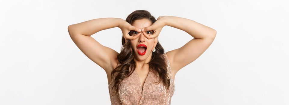 Concept of New Year celebration and winter holidays. Close-up of amazed and excited woman in luxury dress, red lipstick, looking through hand glasses, white background.