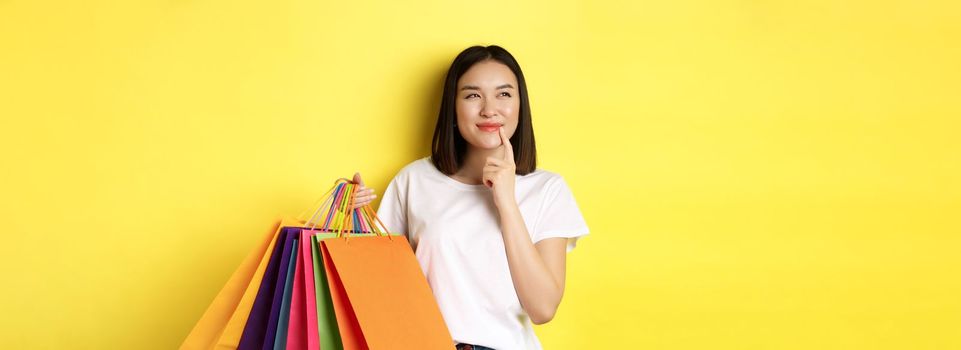 Pensive young woman smiling intrigued, showing shopping bags, thinking about buying something, standing over yellow background.