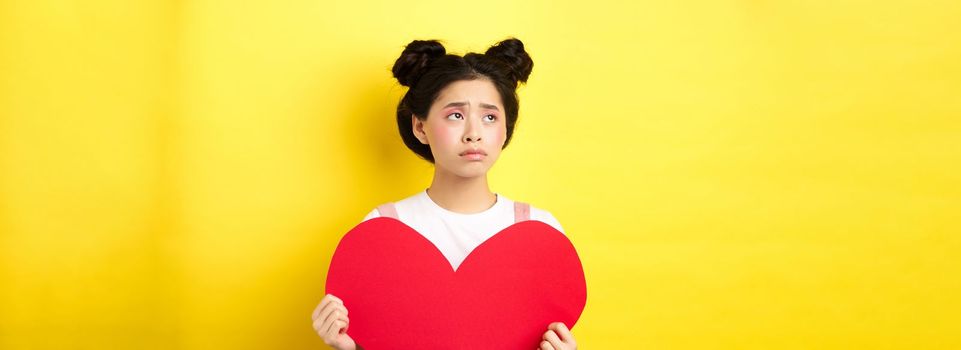 Sad and lonely asian girl standing in make-up with big red heart cutout, left alone on Valentines day, looking left disappointed or upset, standing on yellow background.