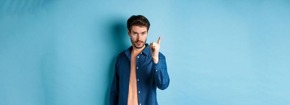 Serious young man scolding person, shaking finger disapprovingly, warn or prohibit something bad, standing on blue background.