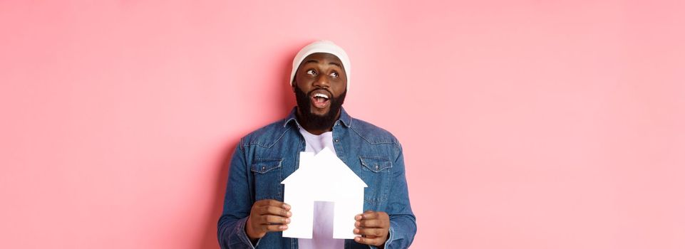 Real estate concept. Handsome african-american man dreaming about home, holding house model and looking at upper left corner, imaging apartment, pink background.