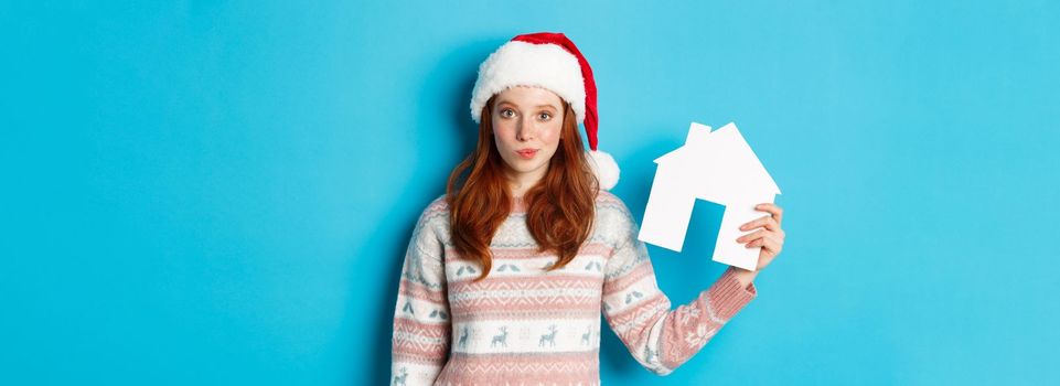 Holiday promos and real estate concept. Cute redhead woman in santa hat and sweater showing paper house model, apartment offer, standing over blue background.