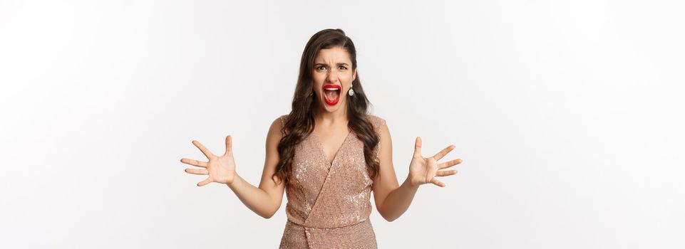 Celebration and party concept. Angry young woman in glamour dress shouting at you, looking mad and outraged, shaking hands aggressive, having an argument, white background.