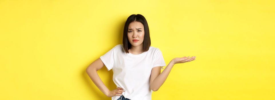 Disappointed asian woman asking so what, raise hand up and staring skeptical at camera, standing over yellow background.