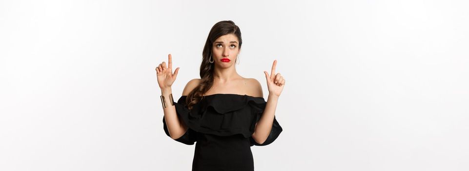 Fashion and beauty. Silly woman in black dress, red lips, looking and pointing fingers up with unamused doubtful expression, white background.