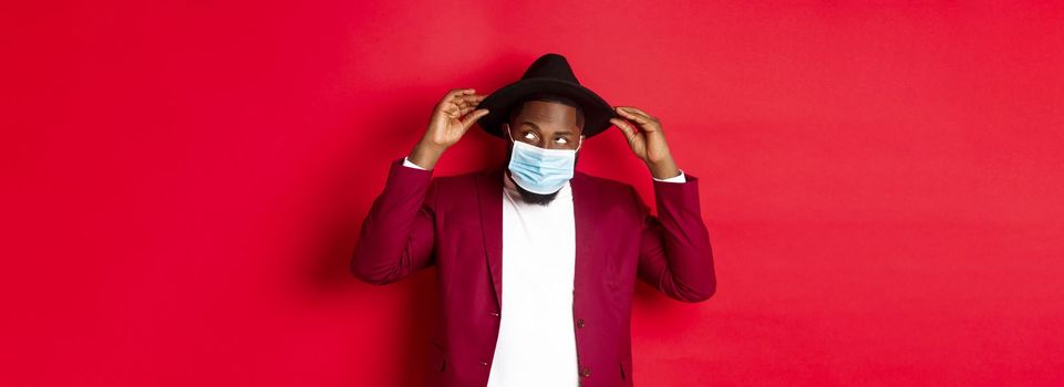 Covid-19, quarantine and holidays concept. Handsome and stylish african american man in face mask, put hat on head and looking sassy, dress-up for party, standing over red background.