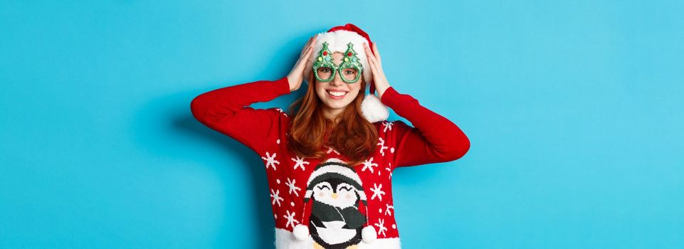 Happy holidays and Christmas concept. Funny redhead teen girl celebrating New Year, wearing santa hat and party glasses, standing against blue background.