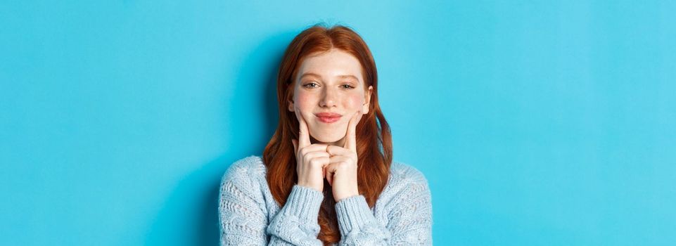 Close-up of cute and silly girl with red hair poking cheeks, showing dimples and smiling, standing against blue background.