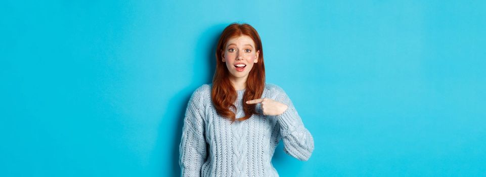 Hopeful redhead girl pointing at herself, standing over blue background.