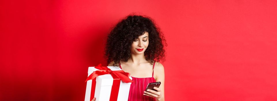Valentines day and shopping. Beautiful caucasian woman in red dress, holding gift box from lover and using mobile phone, reading message on smartphone, studio background.