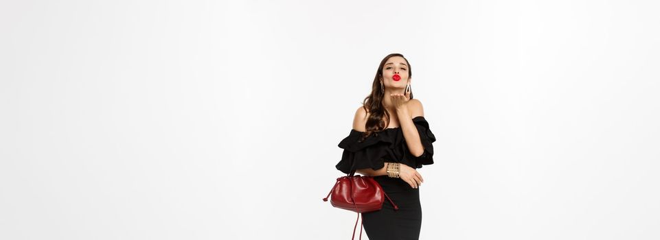 Beauty and fashion concept. Full length of glamour woman with red lipstick and makeup, wearing black dress with purse, sending air kiss at camera, white background.