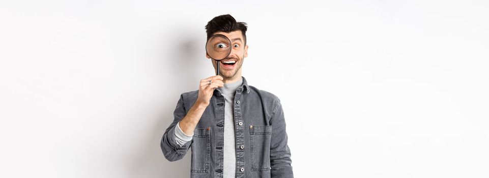 Happy funny guy look through magnifying glass with big eye, checking out interesting deal, searching something, standing on white background.