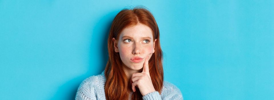 Headshot of troubled teenage girl thinking, looking bothered and frowning, standing against blue background.