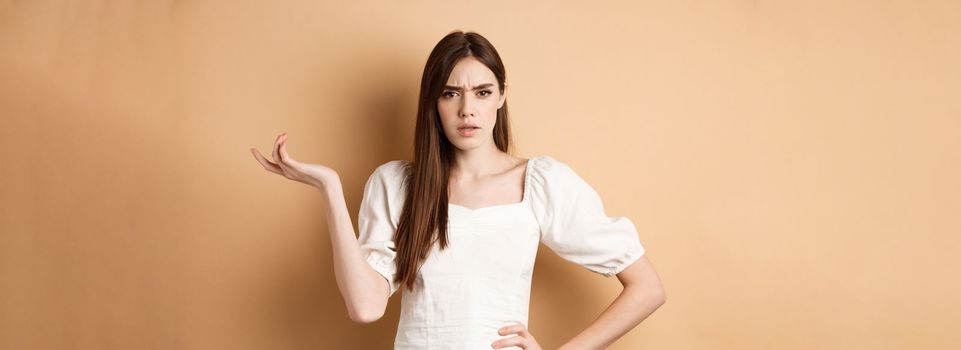 So what. Confused and annoyed young woman raising hand up and staring bothered at camera, cant understand wtf going on, standing on beige background.