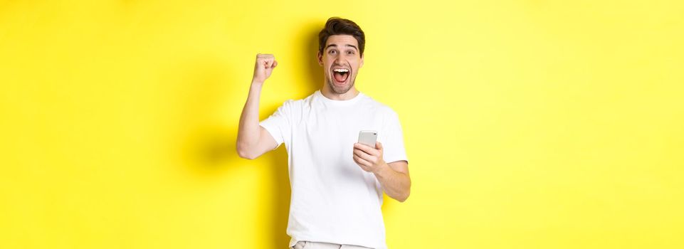 Cheerful man winning on smartphone, raising hand up and holding mobile, achieve app goal, standing over yellow background.