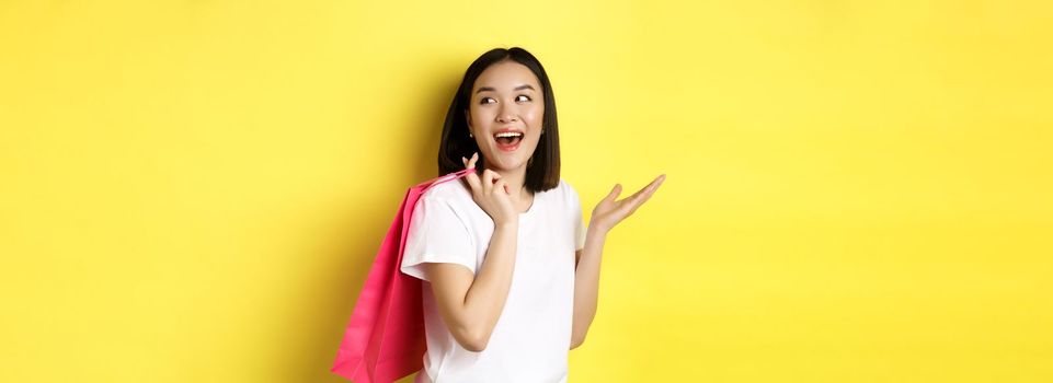 Cheerful asian female shopper looking amused, holding shopping bag and looking right, standing over yellow background.